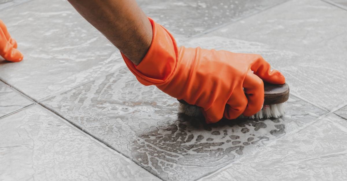 The gloved hand of a homeowner scrubbing his tile bathroom floor with sudsy soap and a bristle brush.