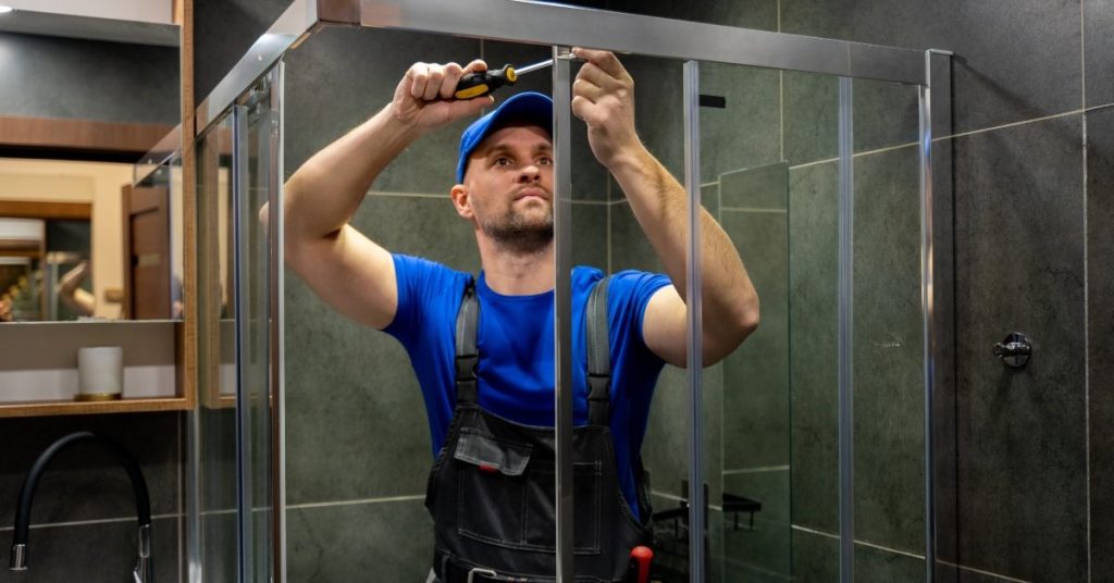 A contractor using a screwdriver to make adjustments to the metal frame surrounding a glass shower door.