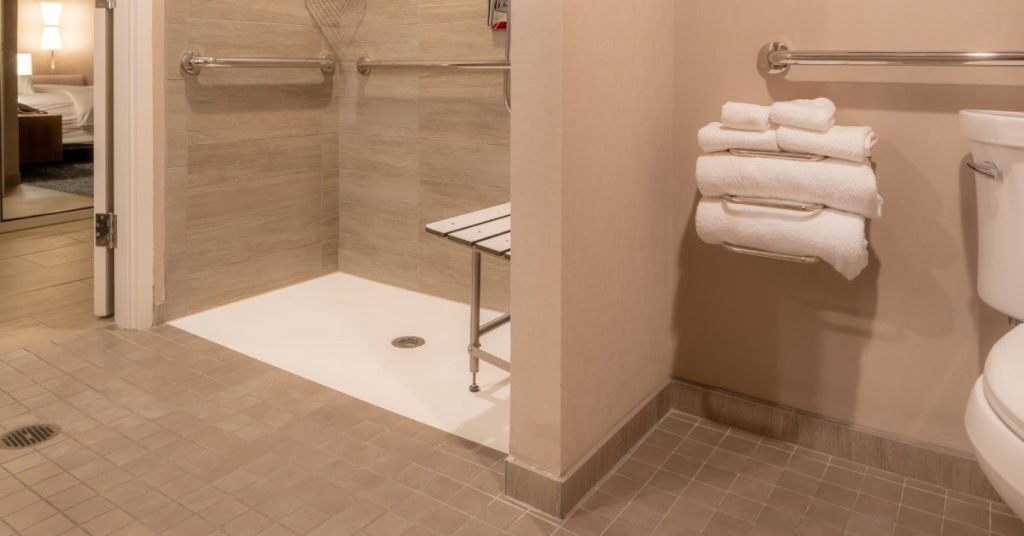 A residential bathroom featuring an accessible, zero-threshold roll-in shower with grab bars and a bench.