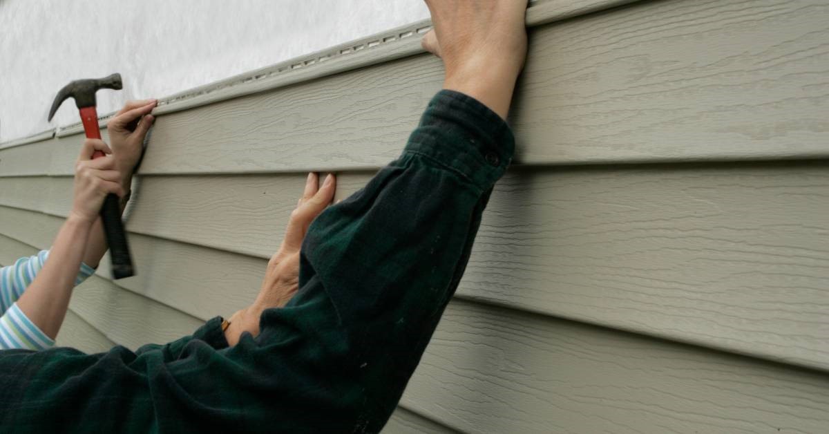 Two people carefully using a hammer and nails to install insulated vinyl siding on the side of a residential structure.