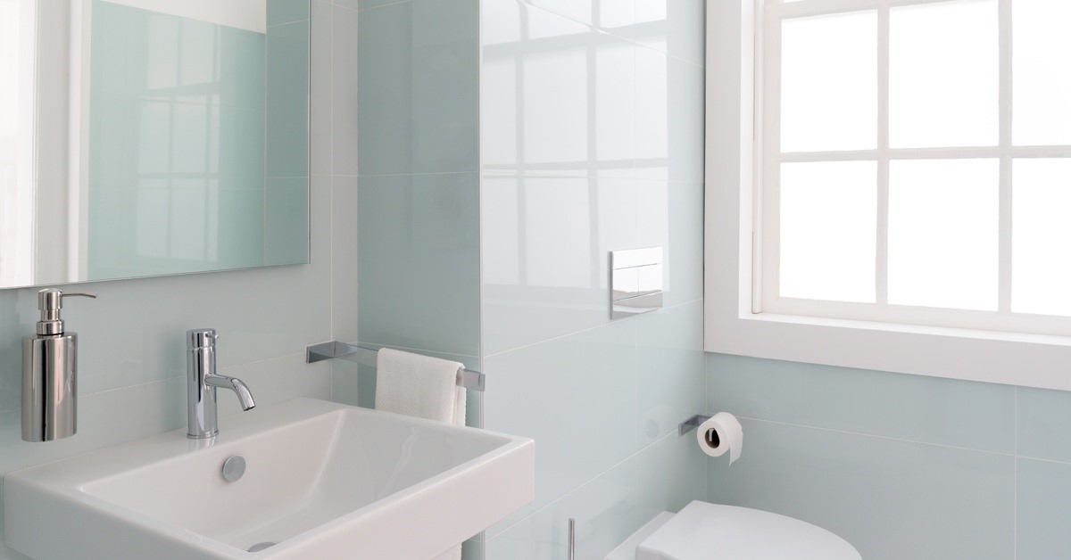A small, modern-style bathroom with light blue walls. The toilet, sink, and window are white and the fixtures are silver.