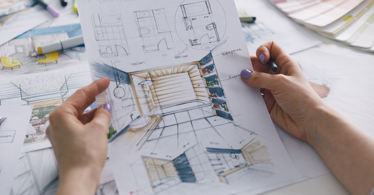 A close-up of a woman holding design plans for a bathroom. Her desk is covered with papers, markers, and paint swatches.
