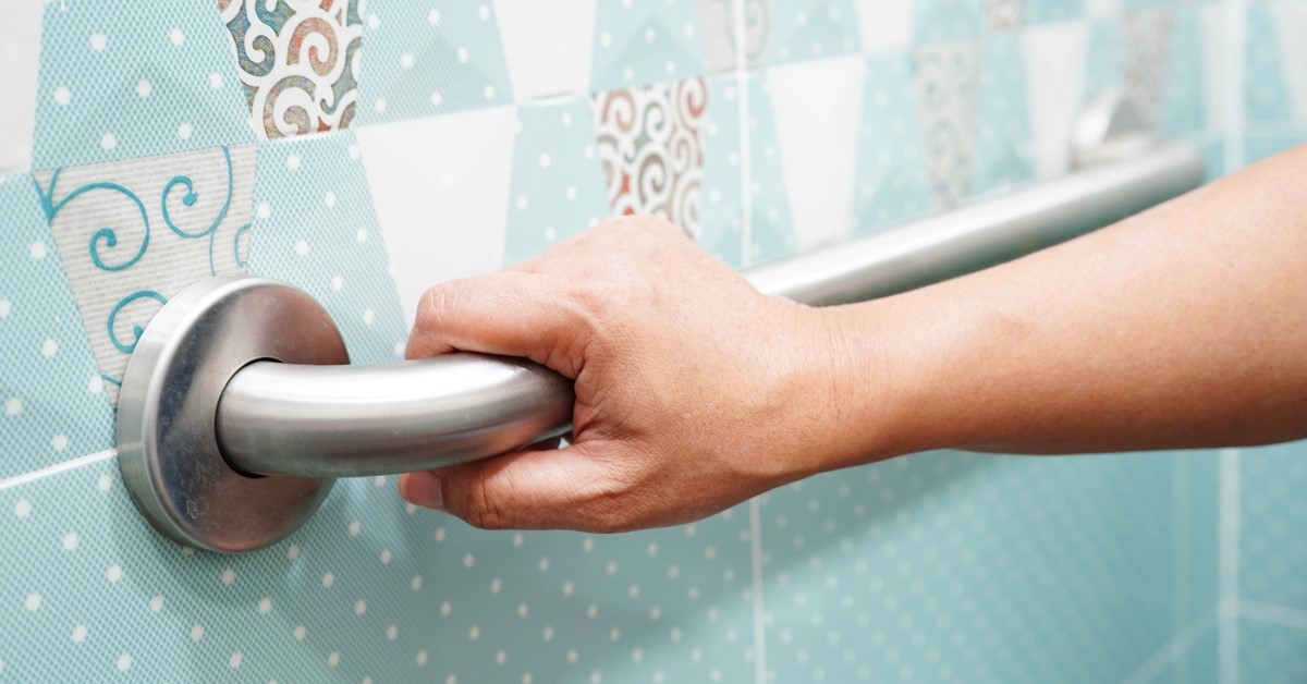 A close-up of a hand grabbing a metal support rail in a bathroom. Behind the rail is blue tile with white polka dots.