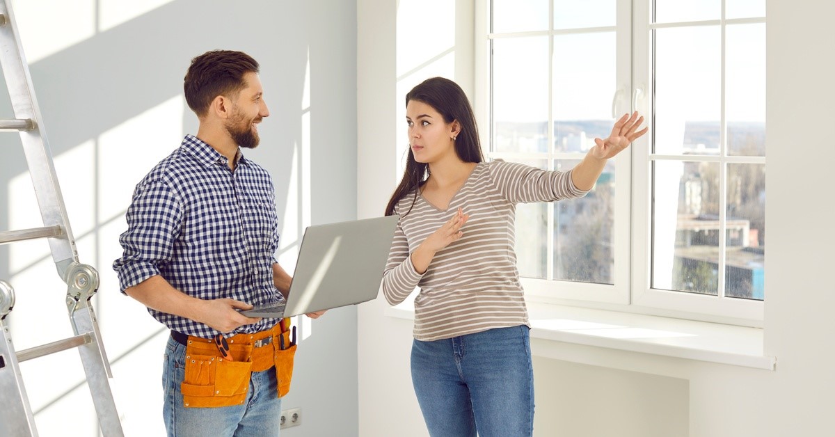 A young woman talking to a contractor who is standing near a ladder while wearing a tool belt and holding a laptop.
