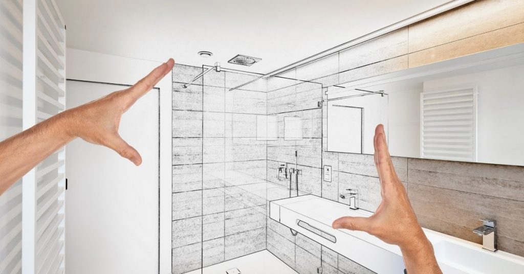 Two hands framing sketched designs for a bathroom renovation, featuring two sinks and a glass shower stall.