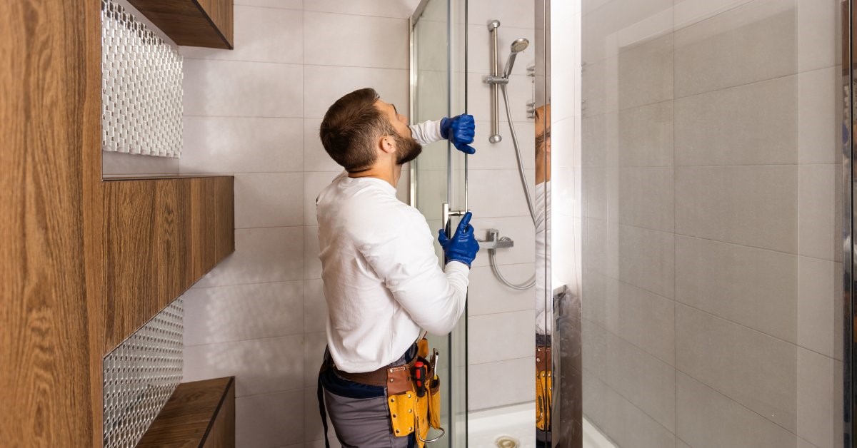 A professional contractor wearing gloves and a tool belt installs the sliding glass door on a new shower.