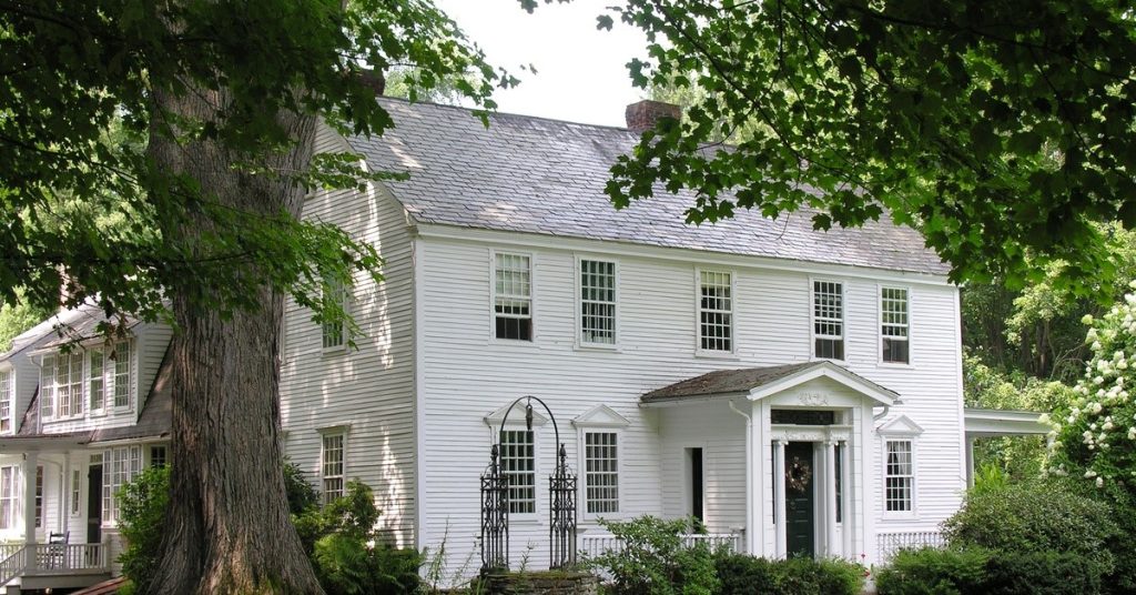 A white New England Colonial-style home surrounded by bushes and tall trees. A gravel path leads to the front door.