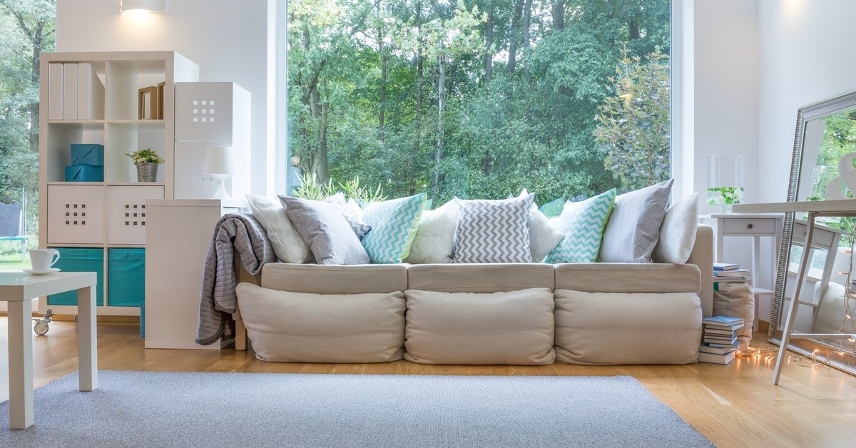 The interior of a living room with a large picture window behind a beige couch with numerous throw pillows.