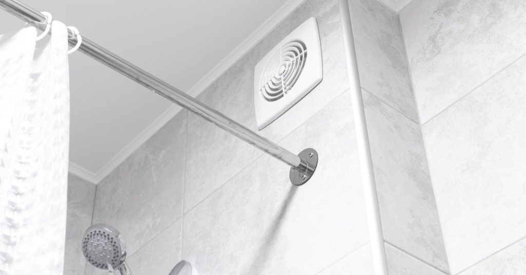 A close-up of a white exhaust fan installed in the gray tiled wall above a showerhead in a bathroom.