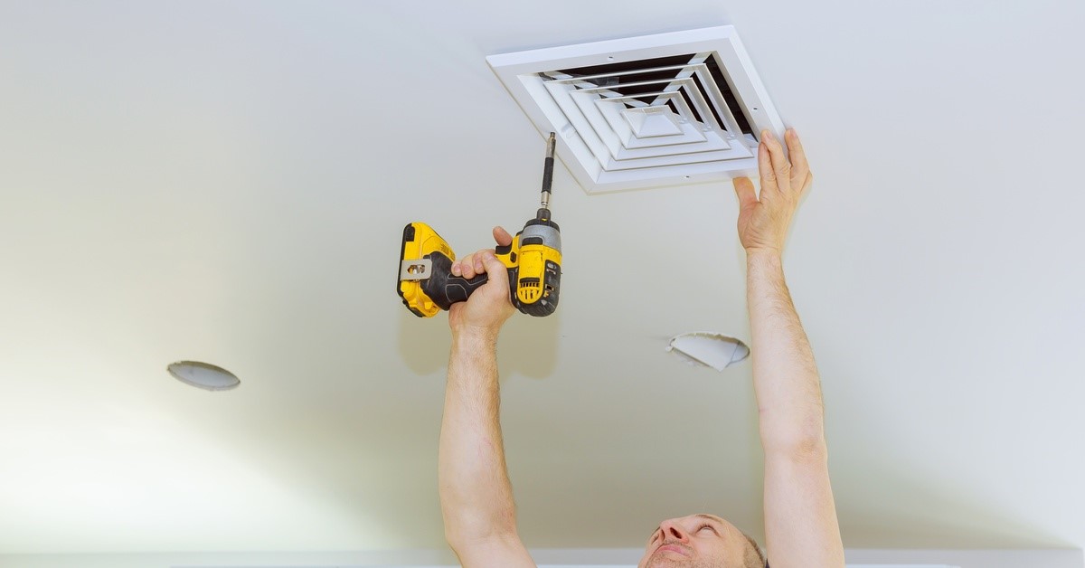 A professional bathroom remodeler using a power drill to install an exhaust fan cover on a bathroom ceiling.