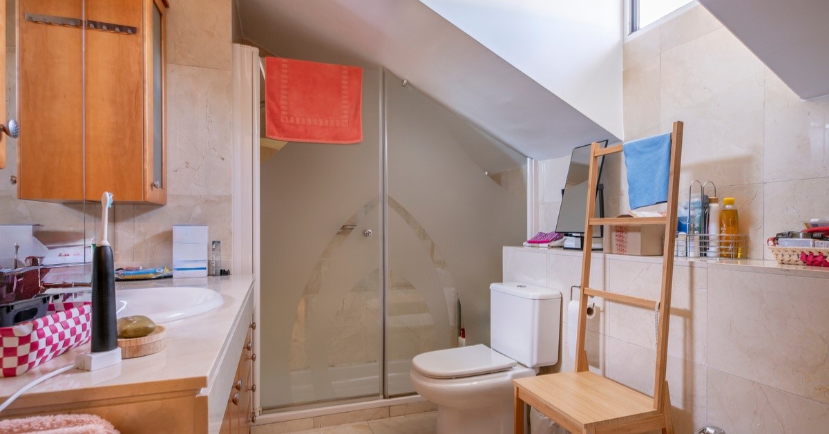 A small bathroom in a home with a slanted ceiling and beige tiled walls. Various toiletries sit near the sink.