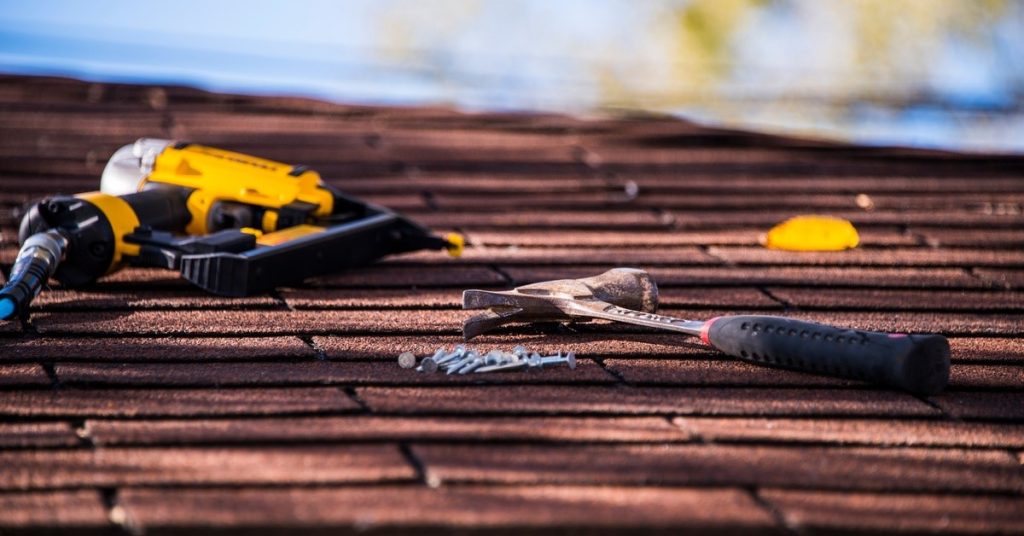 A hammer, a yellow nail gun, and a few nails resting on the surface of a roof constructed of shingles.
