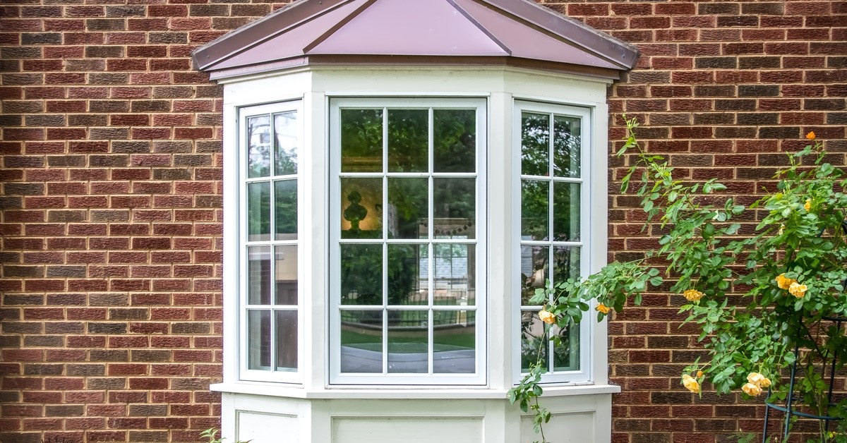 An exterior view of home’s bay window, featuring painted white panes and a copper roof, installed above a garden.