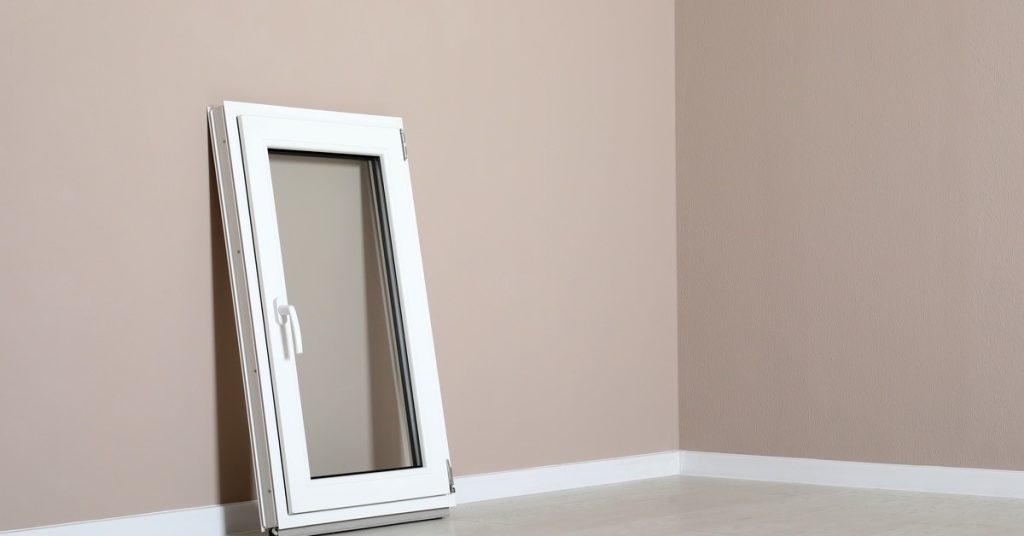 A white, modern single casement window with a handle on its left side leans against a neutral-colored wall and a floor.