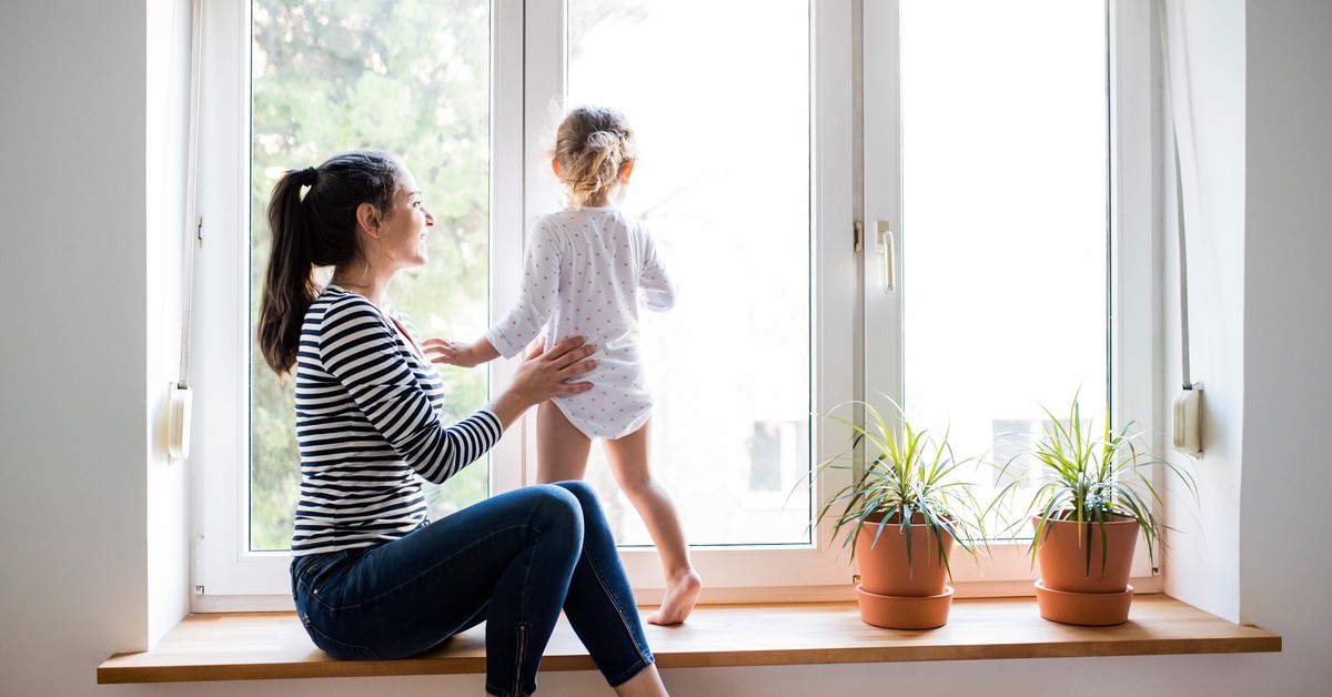 An adult woman sits by a casement window, and a young child stands in front of it as sunlight shines through.