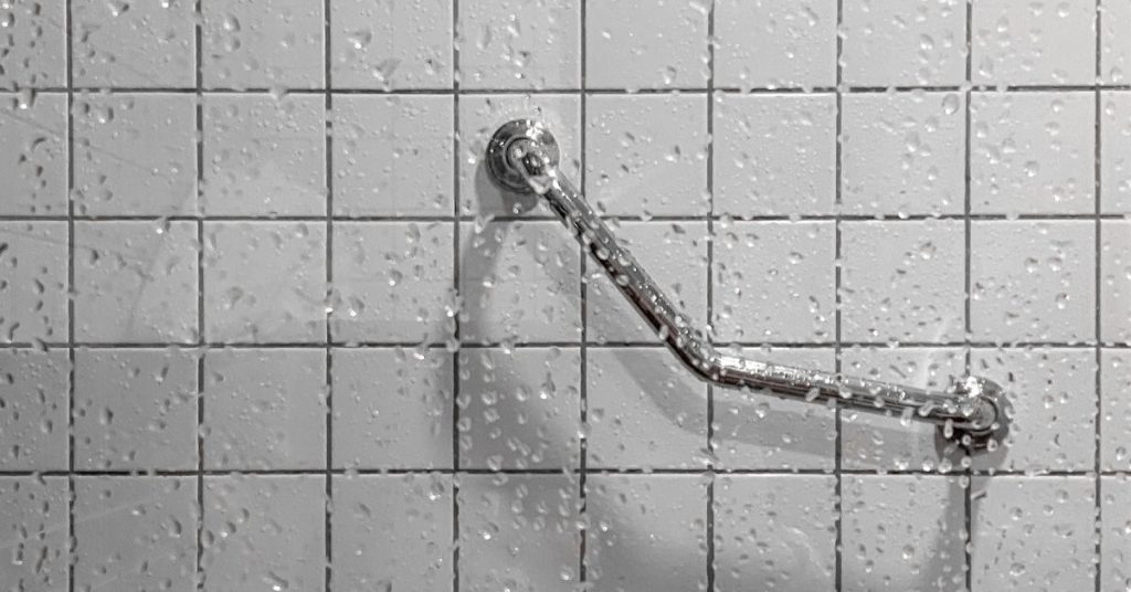 A metal grab bar affixed to the surface of a tiled white shower wall. Water droplets cover the surface.