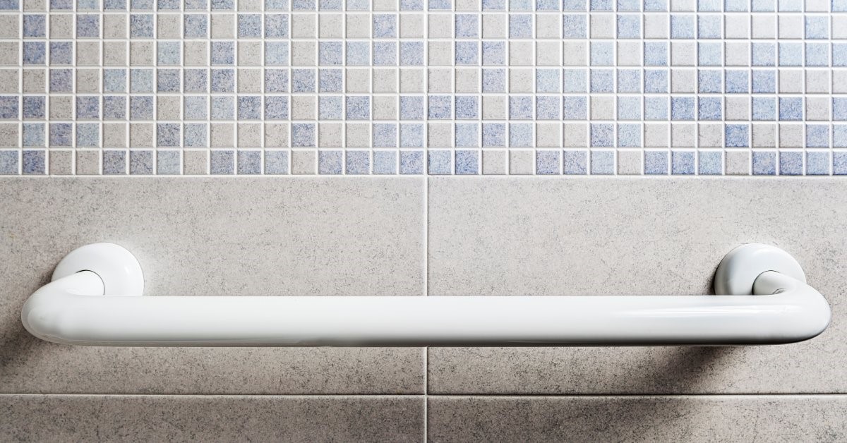 A white grab bar installed horizontally onto a bathroom wall constructed with blue and light-colored mosaic tiling.