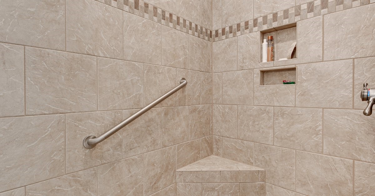 A walk-in shower that includes a safety grab bar, a built-in bench, and a recessed shelf in a beige bathroom.