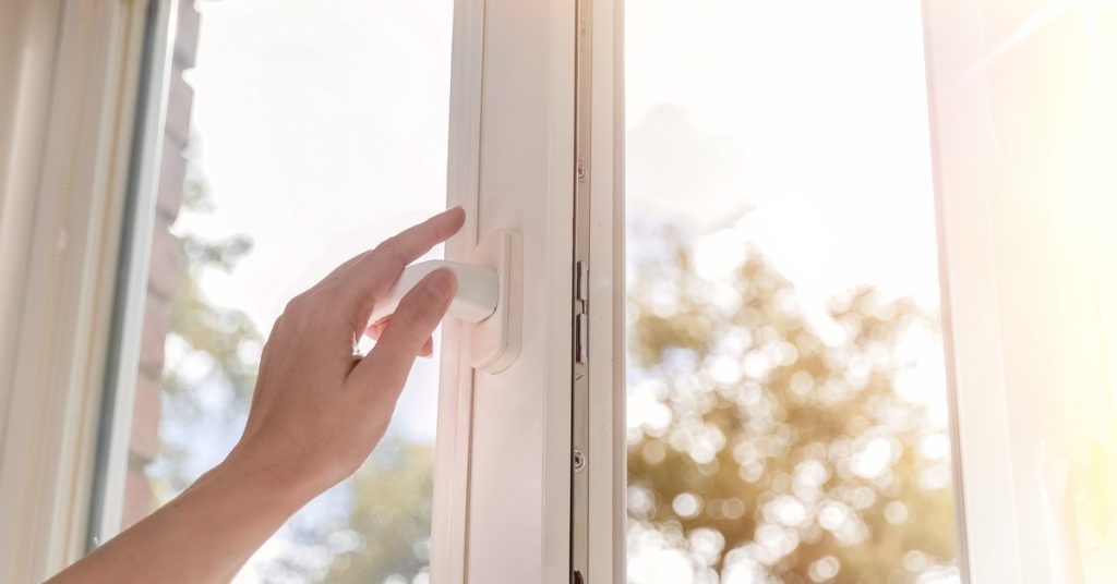 A person's left hand touching the handle of a white window. Sunlight and trees are visible through the window.