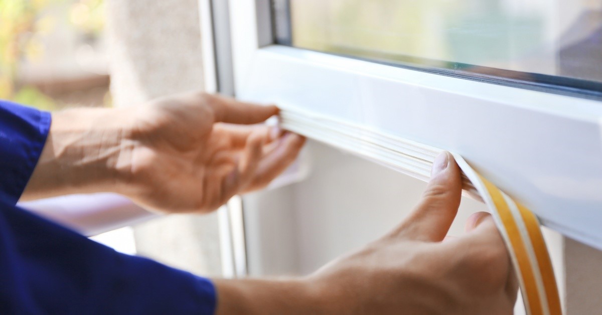 A close-up of a person's hands pressing down on sealing foam tape they affix to the white frame of a window.