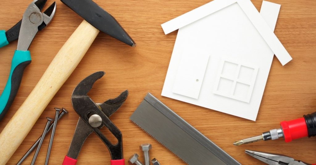 A house-shaped cutout made of white paper on a wooden surface surrounded by tools used in building and repairs.