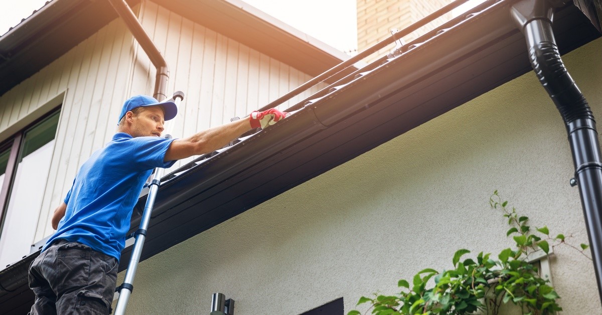 A man wearing a blue shirt and a blue cap stands up on a ladder and removes leaves and dirt from a home's gutters.