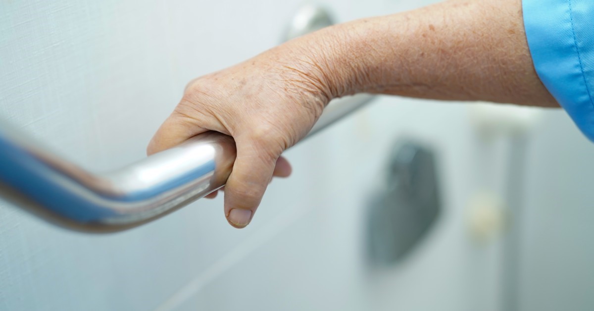 A person uses their right hand to grip the surface of a metal grab bar installed on the wall of a bathroom.