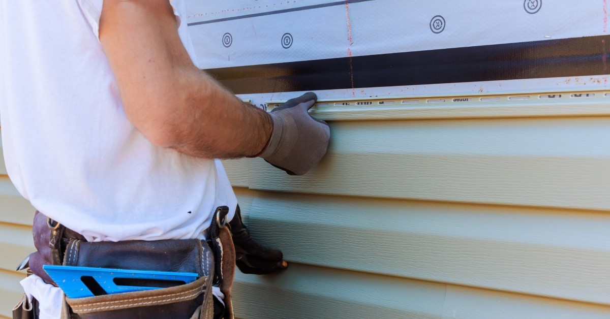 A person wearing a tool belt and gloves installs vinyl siding during the construction of a property.