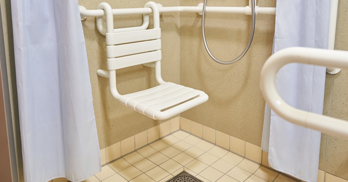 A bathroom with a roll-in shower with tiled walls and flooring, a chair against the wall, and light blue curtains.