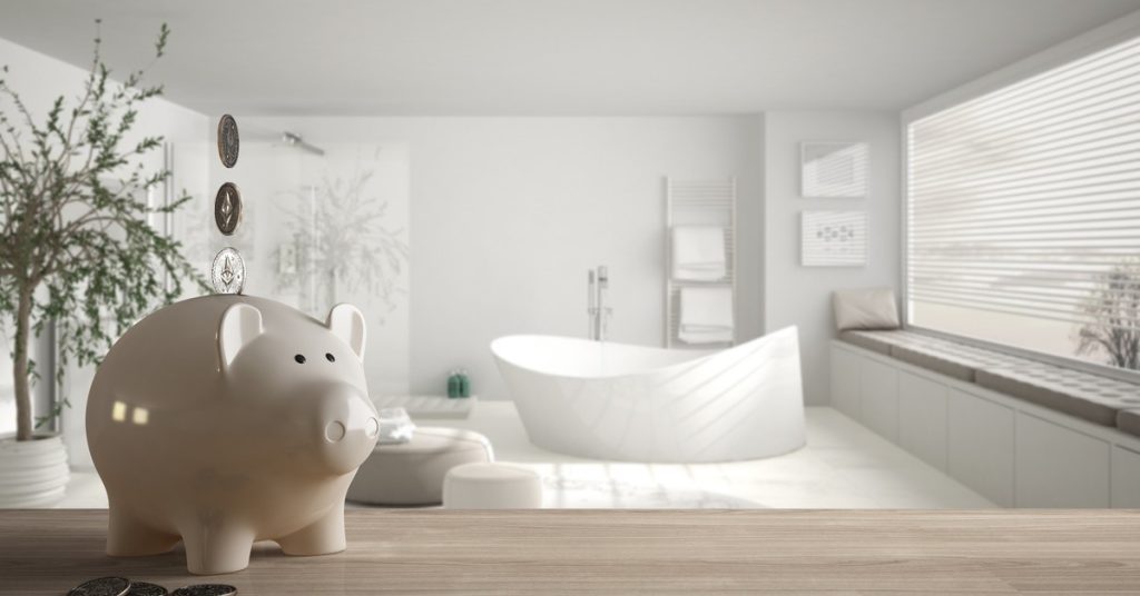 Coins stacked above a white piggy bank sitting on a wooden surface with a bathtub and window in the background.