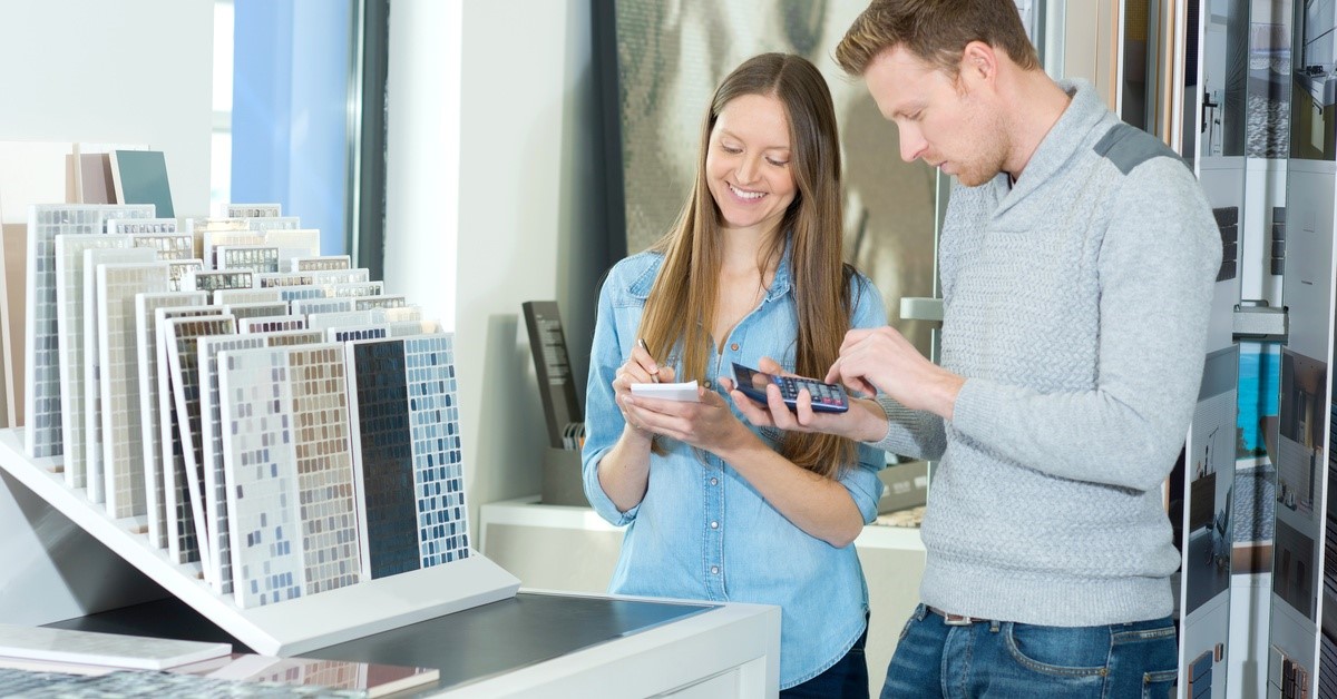 Two people standing together in front of tiling samples. One person holds a pen and notepad, and the other uses a calculator.