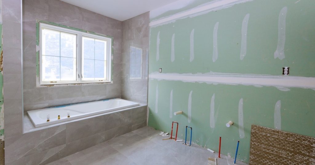 A bathroom with a tub and two windows on one side, and work tools and a green wall with white paint on it on the other side.