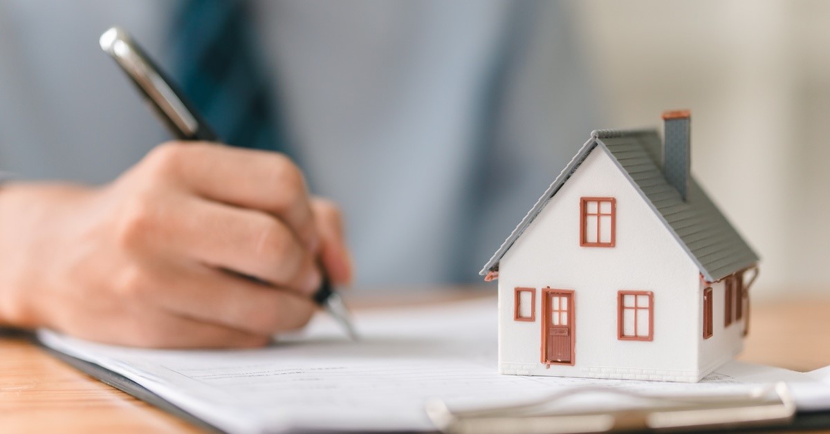 A person holds a pen in their right hand and presses it against a document. A miniature house sits on top of the document.