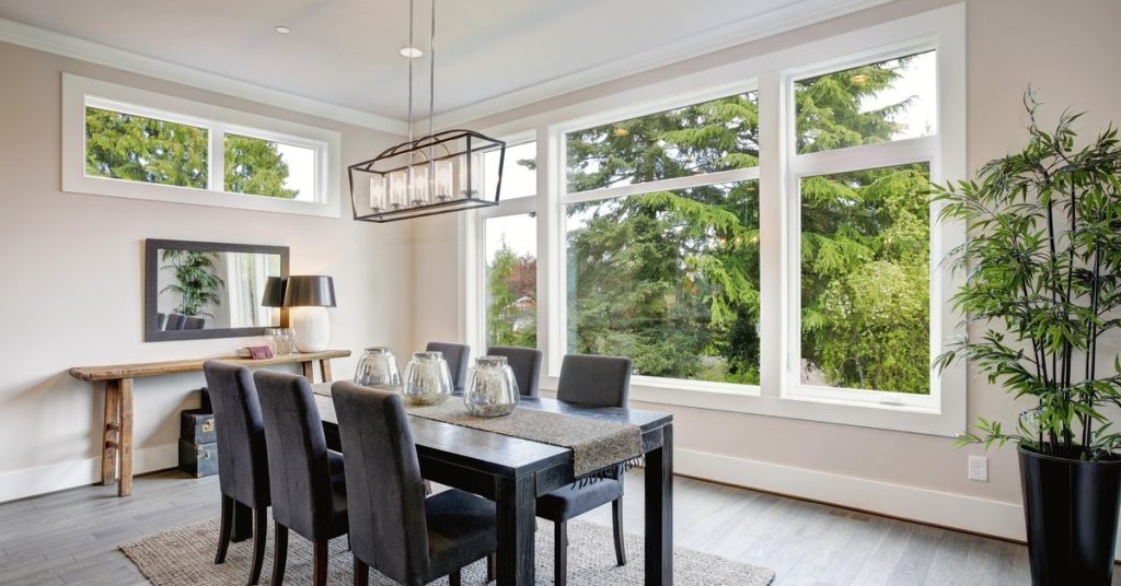 A dining room with a large picture window. There are plants inside the room as well as greenery outside the window