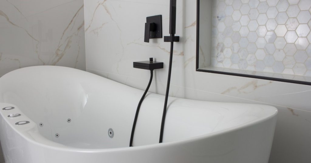 A freestanding bath tun is next to a white tile wall. There is a shower head installed next to the tub.