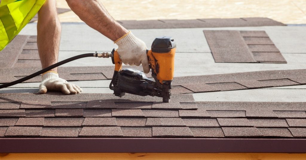A man is holding a nail gun. He’s pointing the nail gun down over some shingles. There are other shingles strewn about.
