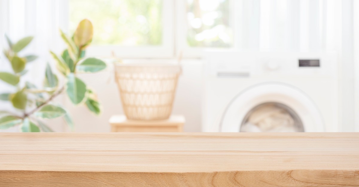 Blurry in the background there is a laundry machine, a basket, and a green plant, while there’s a wooden ledge in the foreground.