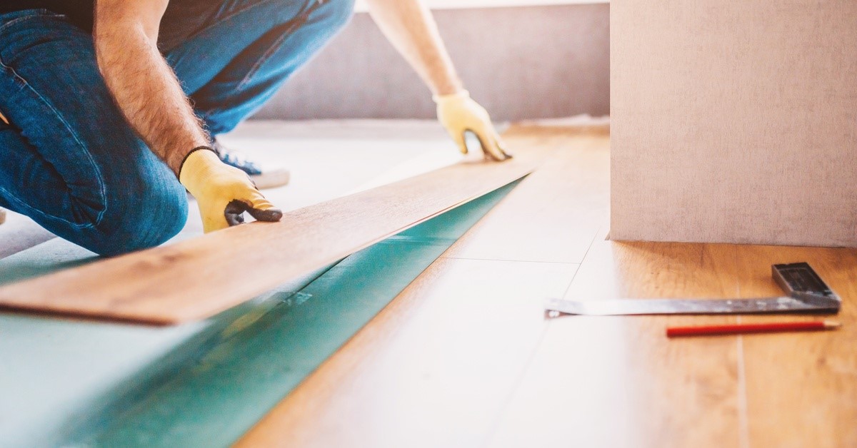 A man wearing yellow gloves is placing a floor tile on the ground. He is lining it up with other pieces already placed.