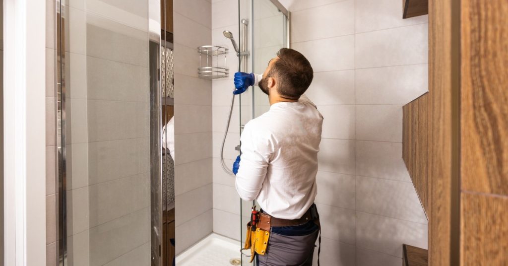 A man with short brown hair is standing next to a shower with glass walls. He is holding the door open.