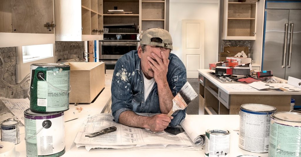 A man in a backwards hat is leaning on a countertop with his hand spressed agsint his face.