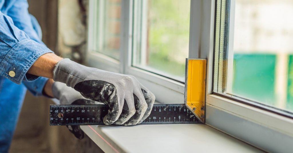 A man in a blue denim shirt is using a tool to see if a window is level and at a 90 degree angle.