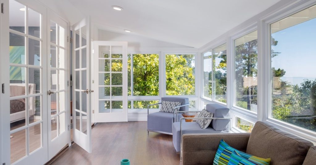 A den area with chairs and windows overlooking a wooded area. There are glass doors leading into the rest of the house.