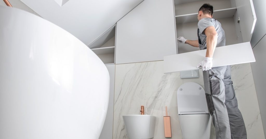 A man is standing in a bathroom while holding a white shelf in his arm. The man is looking at an open cabinet.