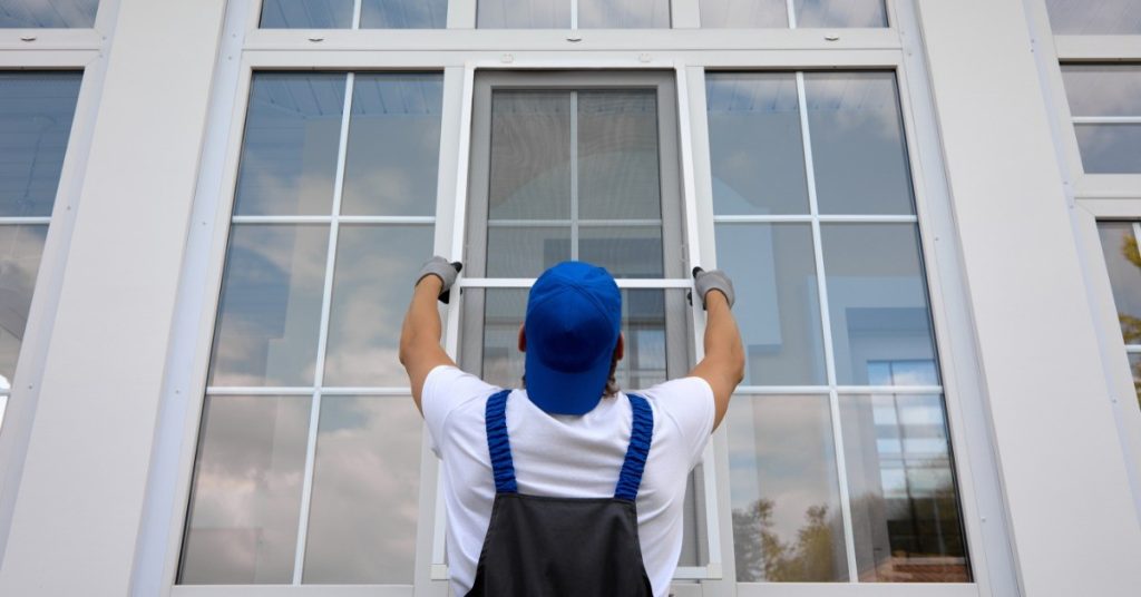 A man in blue overalls is placing a window in a frame. There are clouds reflected in the window panes.