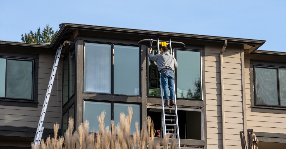 A man in jeans and a gray jacket is standing on a ladder that is propped up against a house.