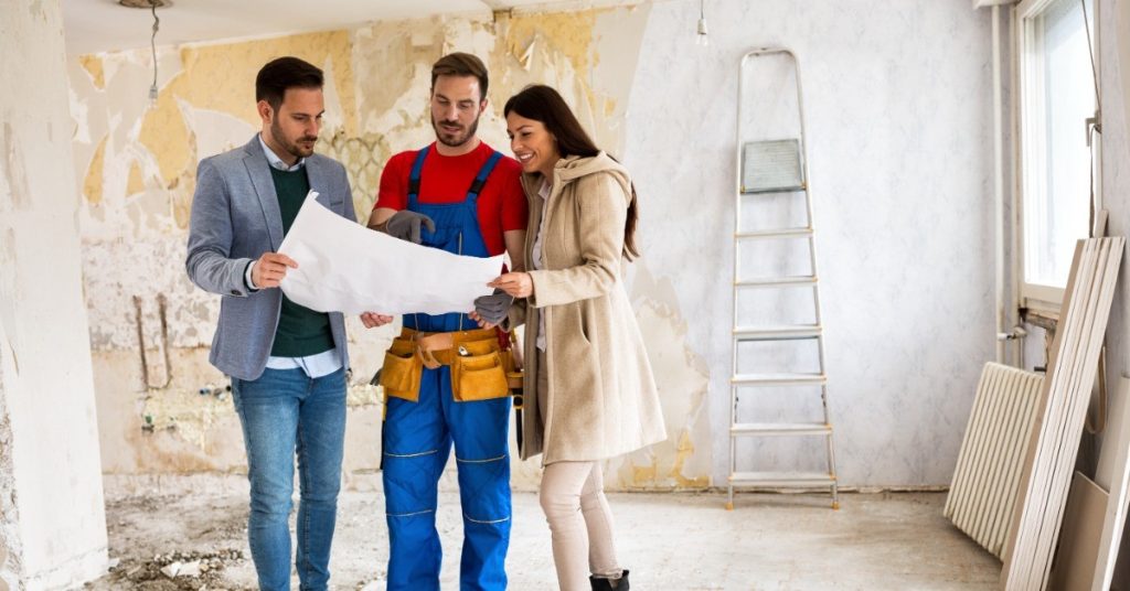 A man and a woman are standing on either side of another man in a red shirt and blue overalls—all are looking at a document.