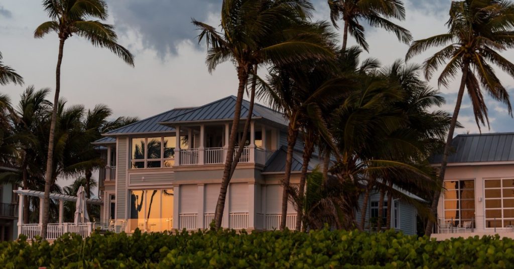 A two-story house has palm trees planted around it. The palm trees are bending in the wind.