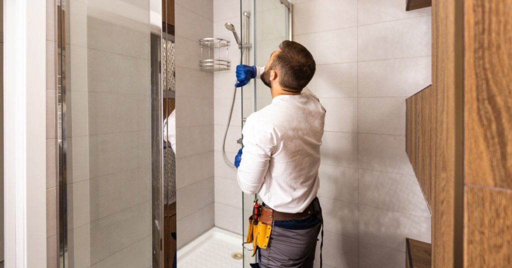 A man with short brown hair is installing a glass sliding door into a shower. The man has a tool belt.